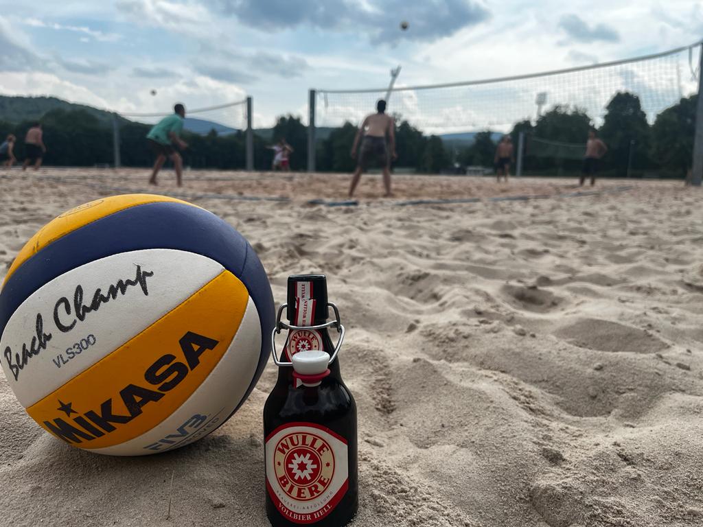 Volleyball und Bierflasche vor dem verschwommenen Hintergrund eines Beachvolleyballfeldes